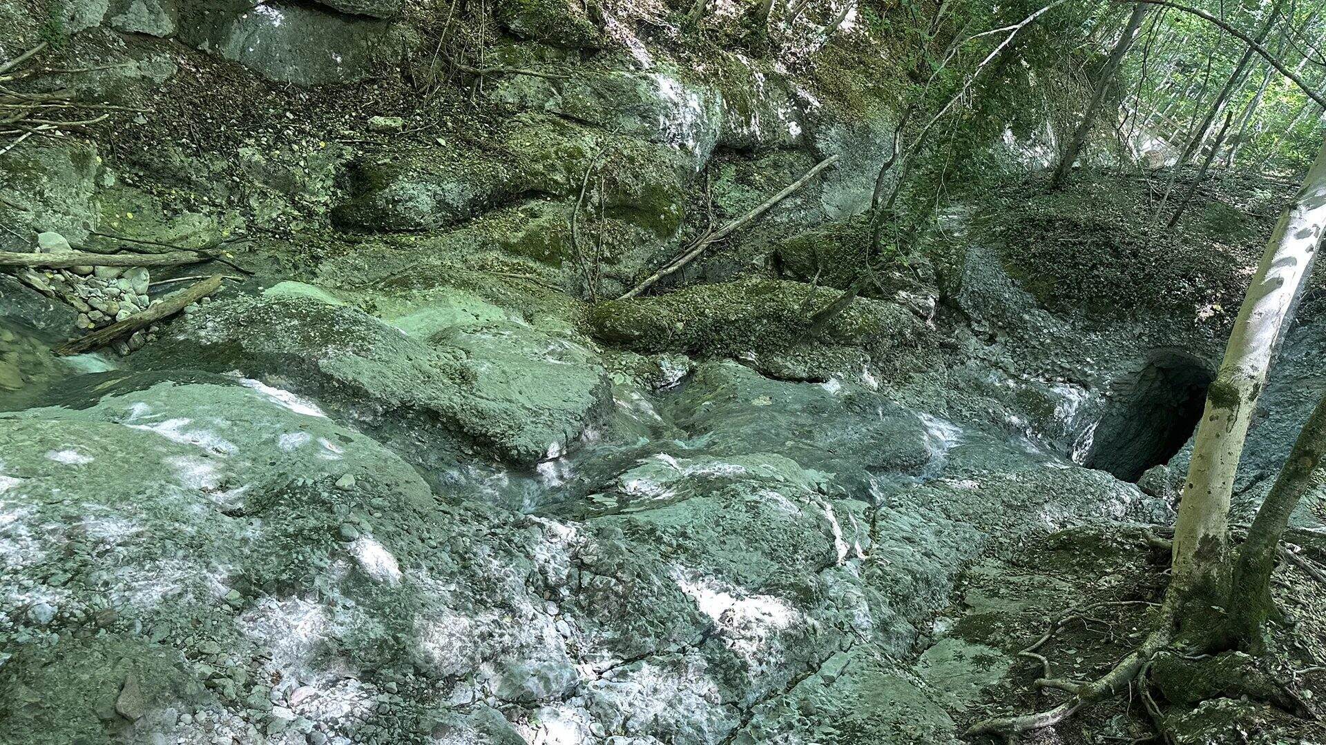 Wasser rinnt über glatte Felsstufen im Bachbett des Aigner Parks, rechts eine dunkle Felsöffnung
